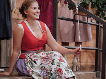Eine Frau sitzt in einem farbenfrohen Dirndl mit Blumenmuster auf einer Holztreppe und hält einen silbernen Ballon an einer Schnur fest. Im Hintergrund hängen Kleidungsstücke an einer Metallstange. | © Gabor Shoes GmbH, Rosenheim