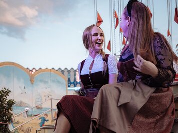 Zwei Frauen sitzen nebeneinander auf einer Treppe vor einem bunten Karussell. Sie tragen festliche Dirndlkleider und Absatzschuhe. Die Stimmung wirkt entspannt und fröhlich. | © Gabor Shoes GmbH, Rosenheim