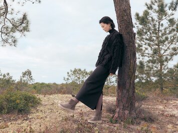 Eine Frau lehnt an einem Baum in einer natürlichen Landschaft und trägt einen schwarzen langen Rock, eine schwarze Jacke und hellbraune Stiefeletten. Der Boden ist von niedriger Vegetation bedeckt, im Hintergrund sind weitere Bäume und ein bewölkter Himmel zu sehen. | © Gabor Shoes GmbH, Rosenheim