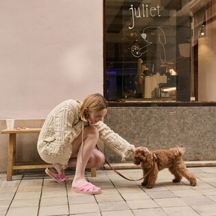 Eine Frau in beigem Strickkleid und rosa Pantoletten kniet auf einem Gehweg und streichelt einen kleinen braunen Hund an der Leine. Im Hintergrund eine Bank und ein Schaufenster mit der Aufschrift "juliet" sowie einer gezeichneten Hundegrafik. | © Gabor Shoes GmbH, Rosenheim