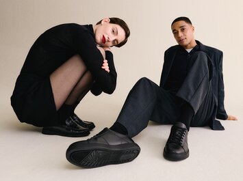 Frau und Mann sitzen entspannt auf dem Boden, beide tragen dunkle, elegante Outfits und schwarze Gabor Schuhe, deren Sohlen im Vordergrund sichtbar sind. | © Gabor Shoes GmbH, Rosenheim